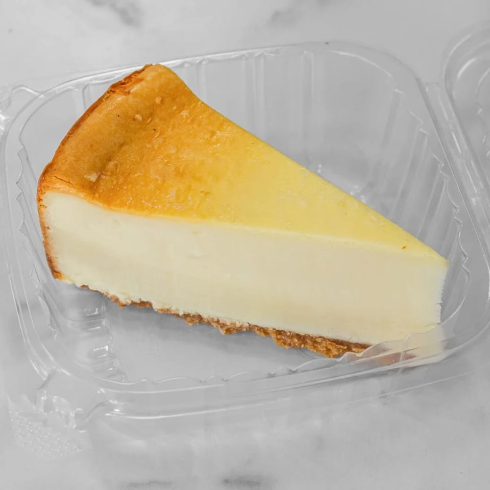 A slice of classic cheesecake with a golden-brown top and graham cracker crust in a clear plastic container.